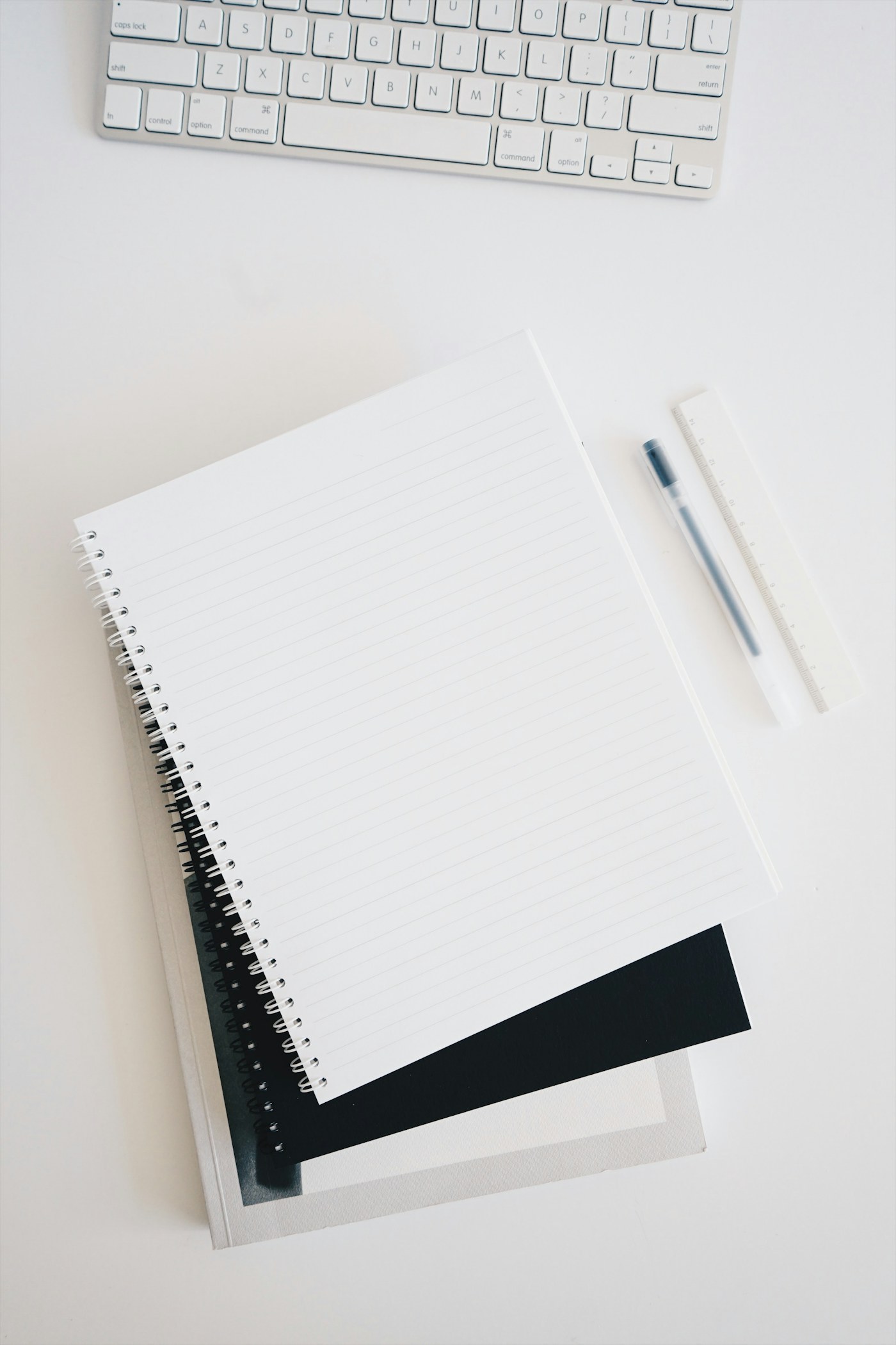 Blank spiral notebook, pen and ruler beside a white keyboard on a pale desk.