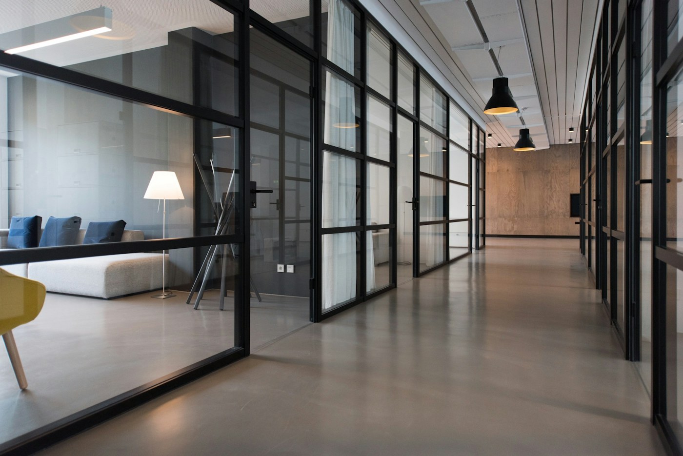 Empty office corridor with glass partitions, lounge seating and pendant lights.